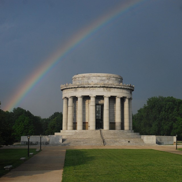 Rainbow over a round memorial with Greek-style columns that has a lawn in front. 