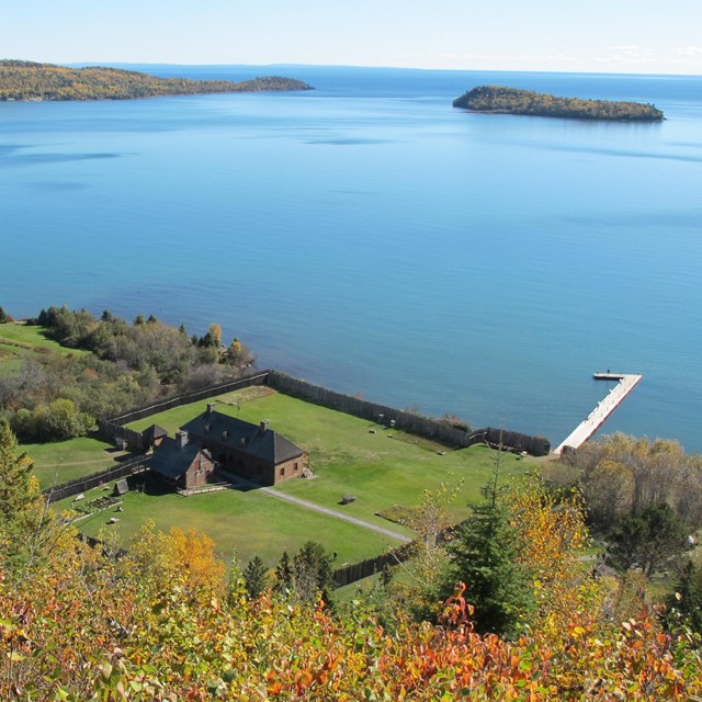 View from a hillside during fall to a building, grounds, and dock that is sticking out into a bay. 