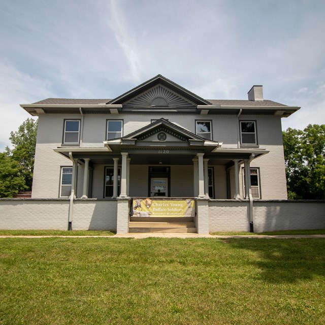 A large grey house with dark grey shutters with a green lawn out front under a blue sky.