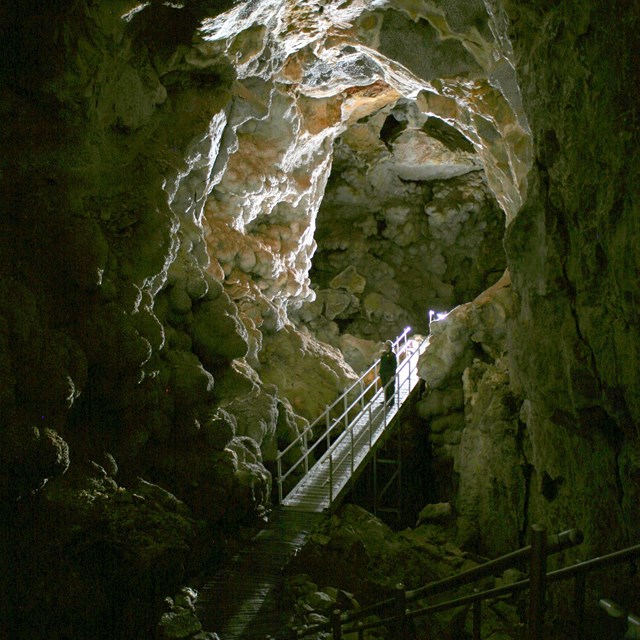 A park ranger stands on a metal platform within a long cave passageway with a vaulted ceiling.