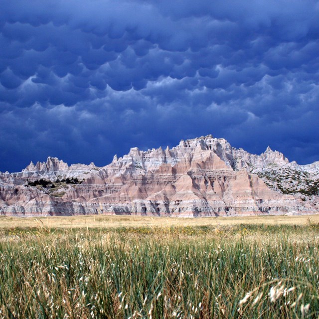 Layered badlands formations behind fields of green grass under cloudy and billowing clouds.