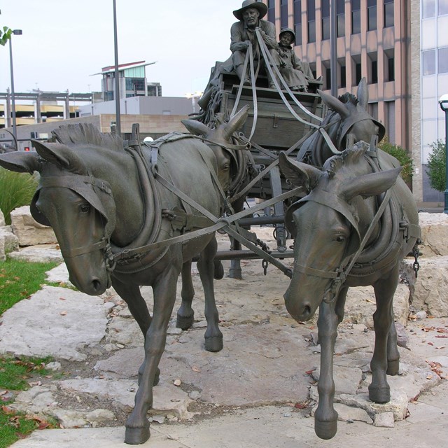 Sculpture of horses pulling a carriage with people sitting on top. Buildings are in the background. 