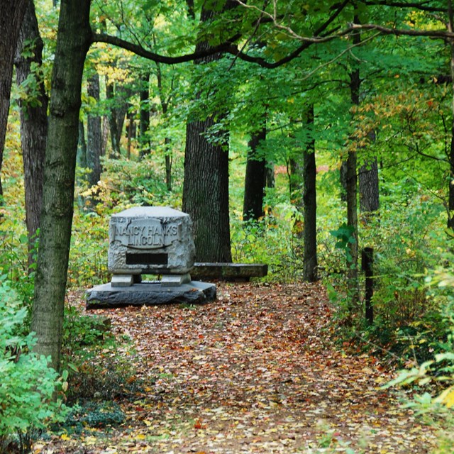 Trail through the woods leading to a stone. Lettering on the stone reads, 