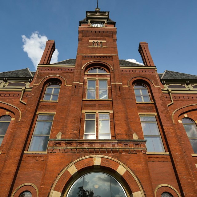 The front of a tall red brick building with a clock tower and many windows. 