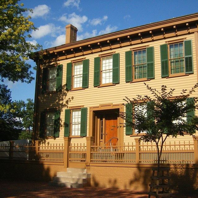 Front of a two story home with yellow trim and green window shutters. 