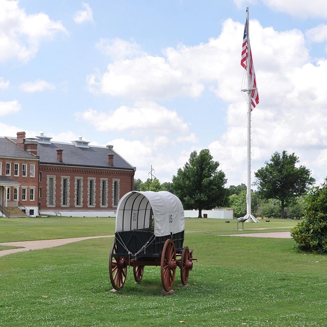 Parade ground with a wagon in front and a large flagpole and stone building behind.