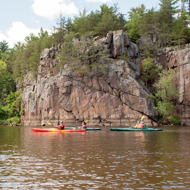 Three kayakers on a river below a cliff face that is lined at the top with trees. 