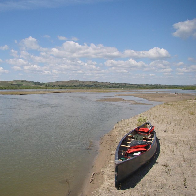 A canoe loaded with gear sits quietly on a sandy river bank with blue sky above.