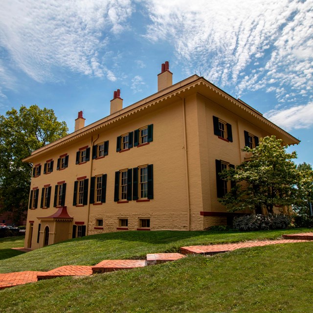 A yellow two-story brick building with tall windows and green shutters on them.