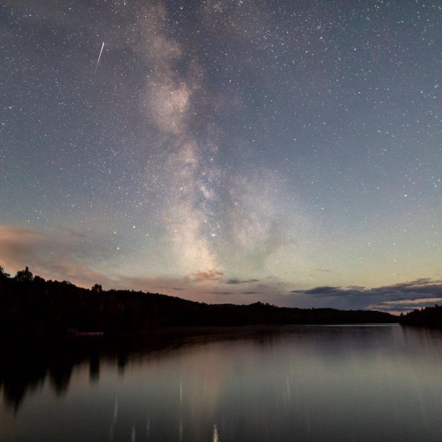 The night sky filled with stars and milky way over water and a forest. 