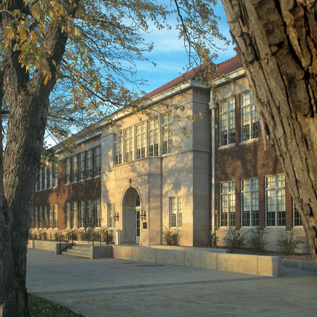 Large two-story grey brick building with trees in front. 