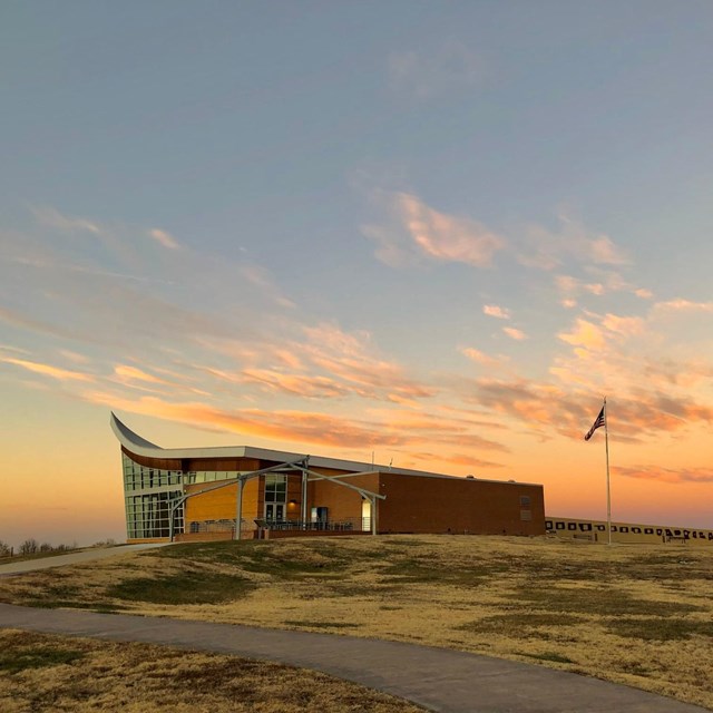 Sunset illuminates the sky above a large building that has a roof that slopes up on one side. 