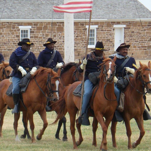 Six Black men dressed in old military uniforms on horseback. One is holding a US flag.