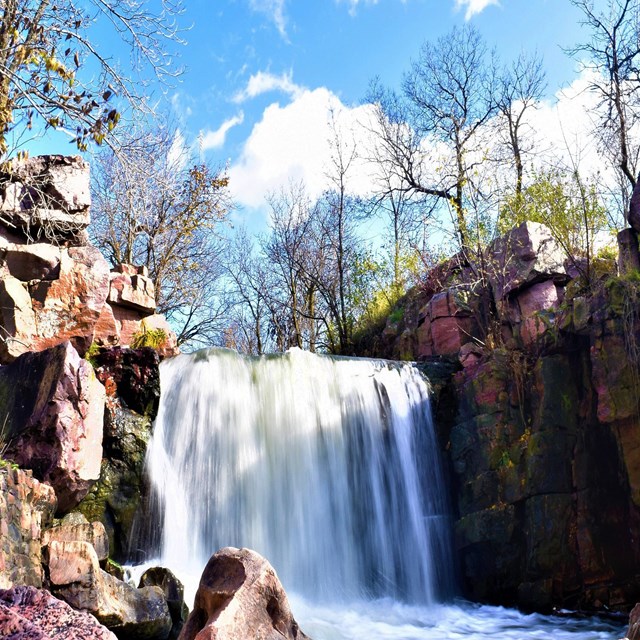 A stream lined with red-colored rocks and deciduous trees goes over a waterfall. 