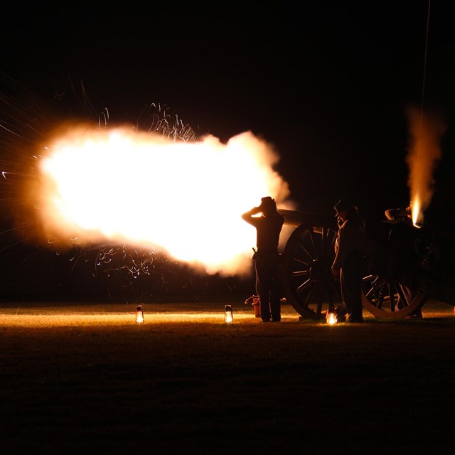 A cannon firing in the dark with people behind. 