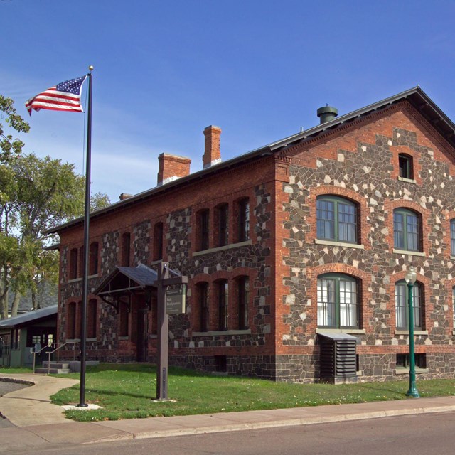 Two story stone and brick building. The American flag flies in front of a blue sky.