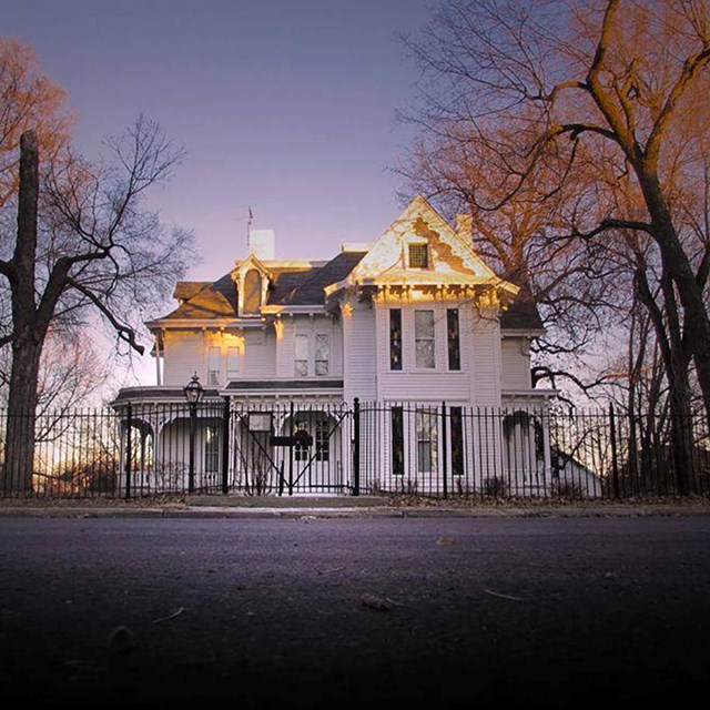 Two story white house with a tall pointed roof with trees on the sides and fence and road in front. 