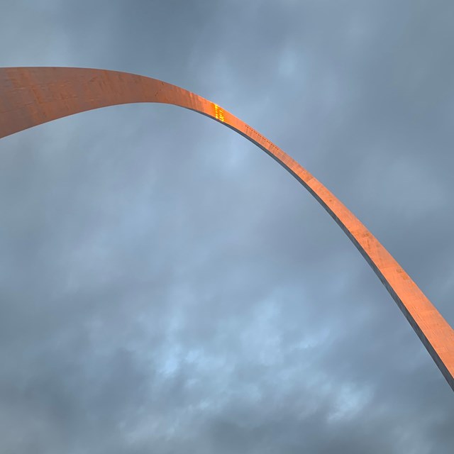Top of a metal arch bathed in orange light during sunrise against a cloudy sky. 