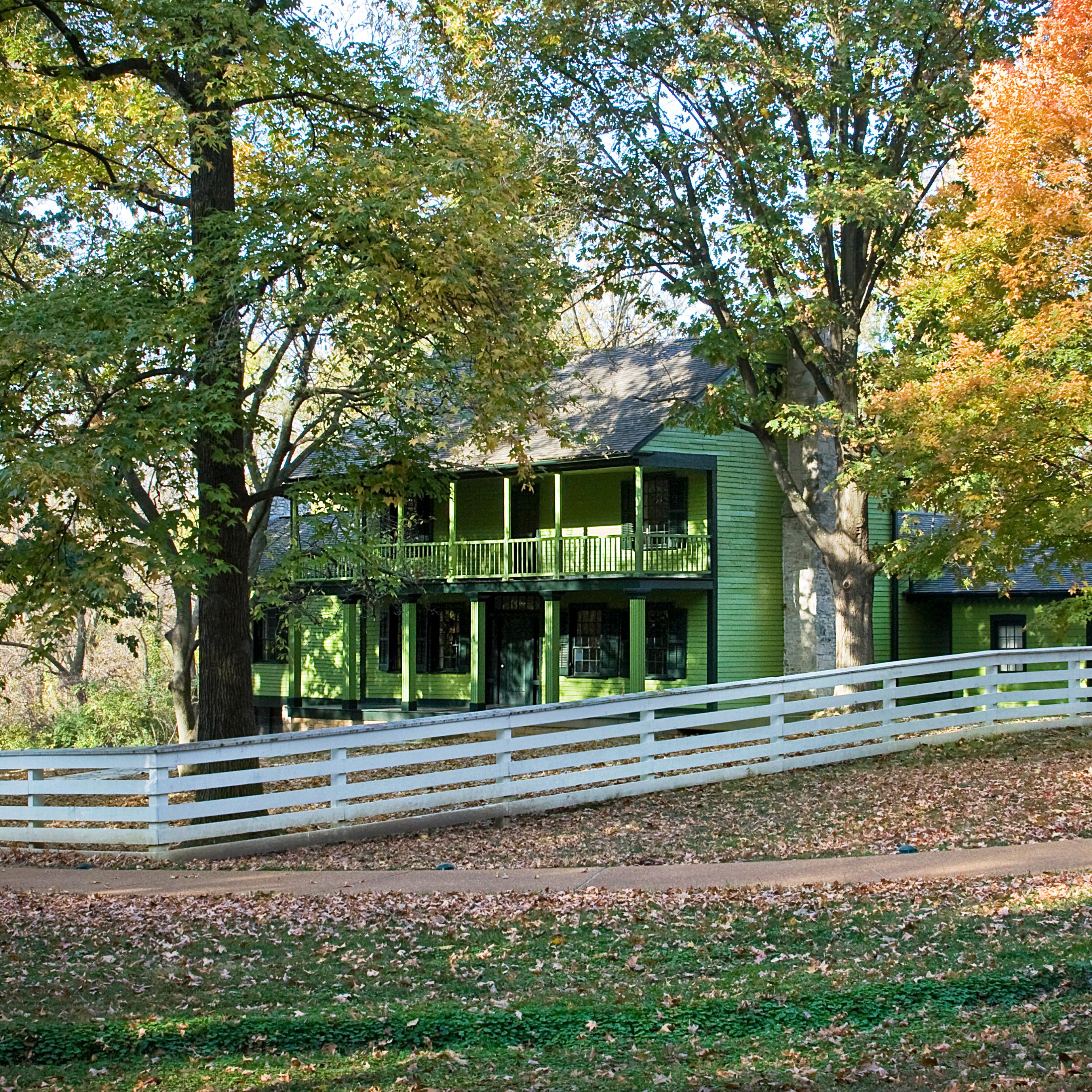 Green house with fence surrounded by trees with green, orange, and yellow leaves.