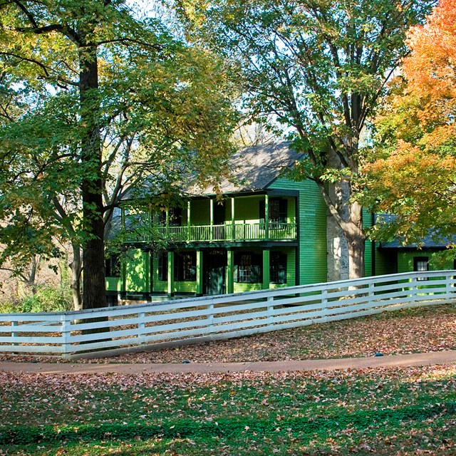 Green house with fence surrounded by trees with green, orange, and yellow leaves.