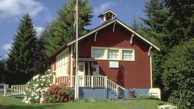 Red Schoolhouse with white trim atop green grass and white flowers.