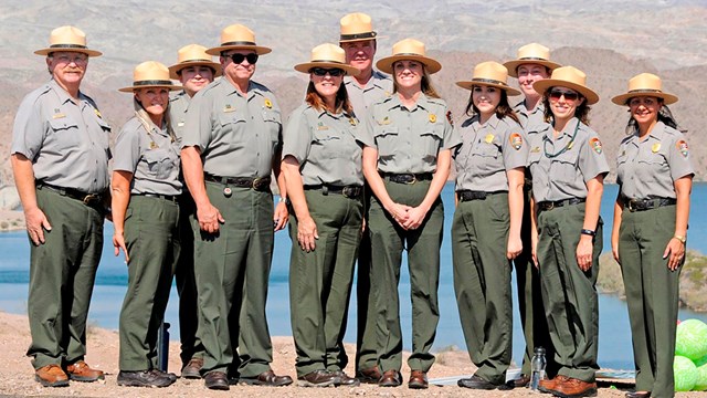Eleven people wear an National Park Service Uniform standing in front of a lake.