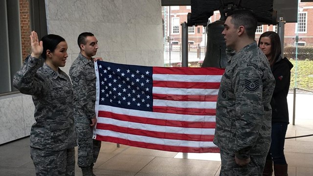 Four people in army uniforms stand at the liberty bell with flag, arms raised for swearing in.