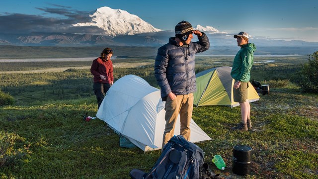 Three people in jackets getting out of a tent. Snow-capped mountains are in the distance.