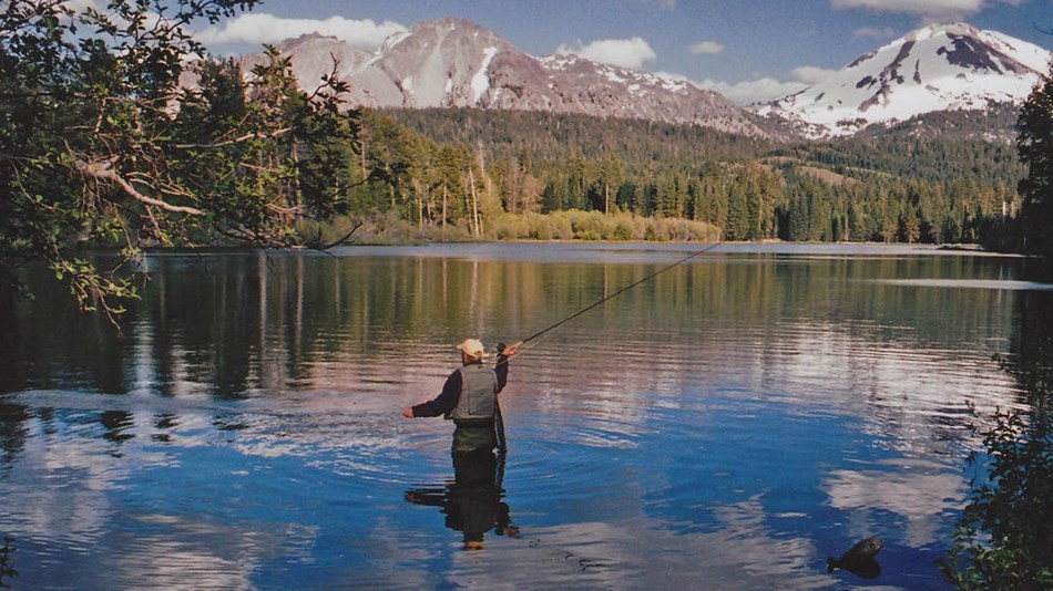 Man wearing fishing vest stands in middle of lake casting his line.