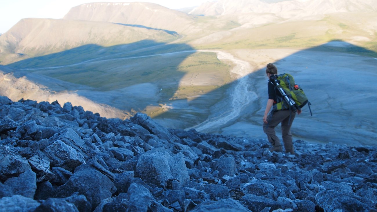 A woman follows a trail down a mountain. Mountains and snow are in the background.