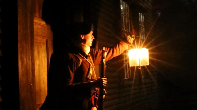 An armed colonial solider stands in the doorway of a wooden house at night holding a lantern