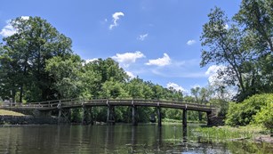 The wooden, Old North Bridge spans the gentle Concord River. Green foliage covers the river banks.