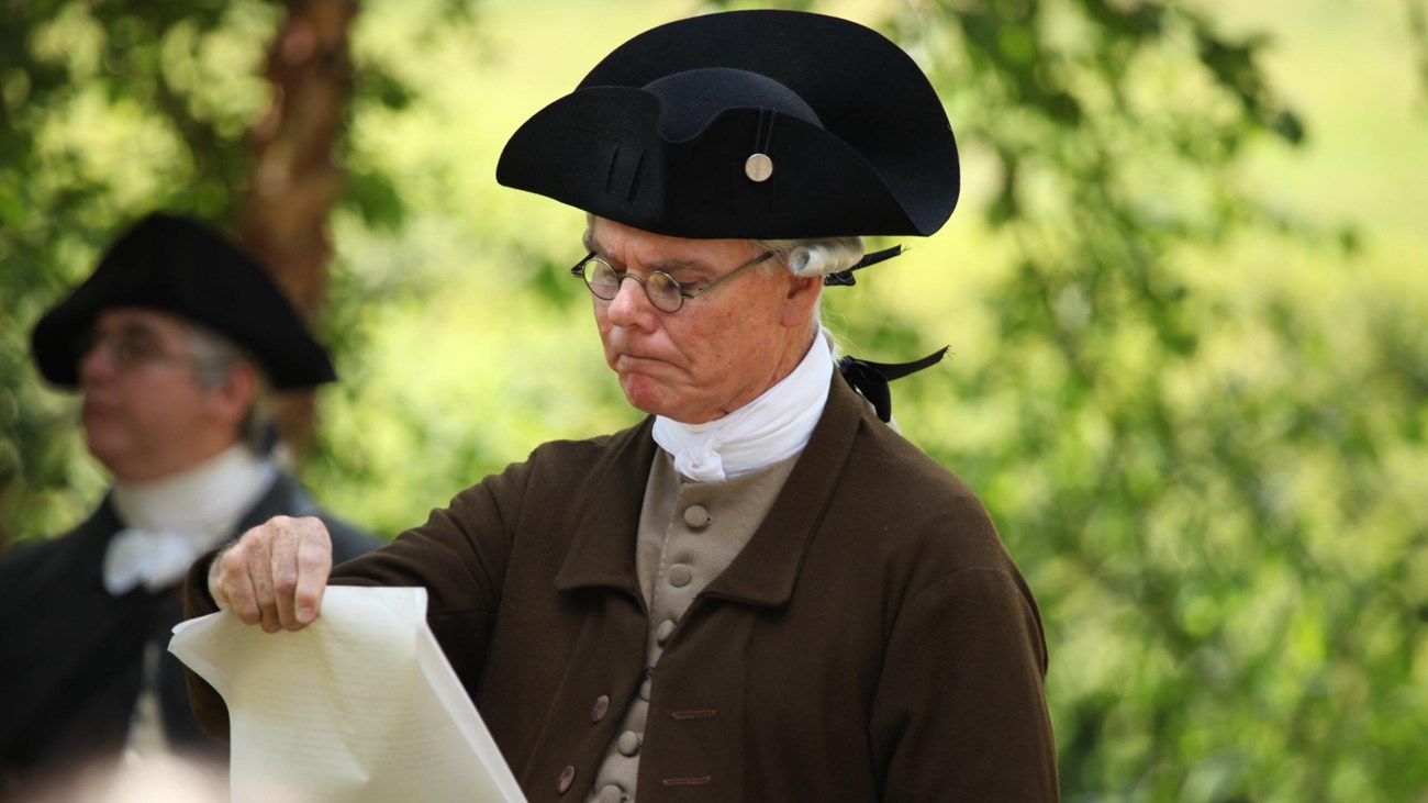 A man in colonial clothing standing outside reads a scroll of parchment. His face is contemplative. 
