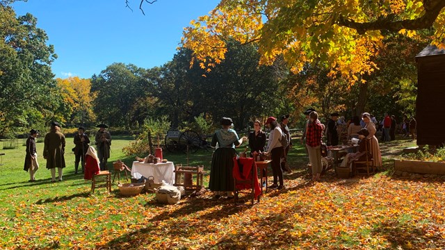 A group of visitors talk with 18th century living historians under trees turning red and orange