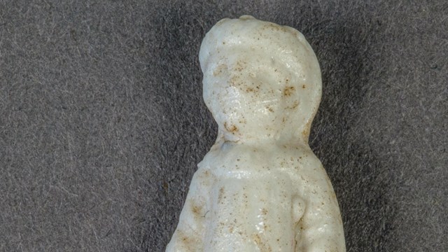A small white ceramic doll with 19th century style clothes and a blank expression