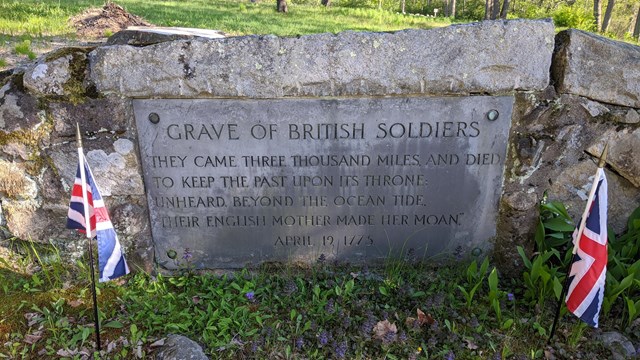 A gravestone is imbedded into a low stonewall surrounded by fencing and green grass.