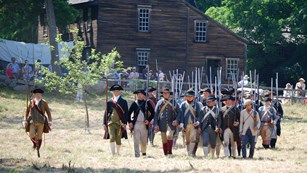 A battle line of Colonial Reenactors march with muskets ready in an open field. Visitors watch 