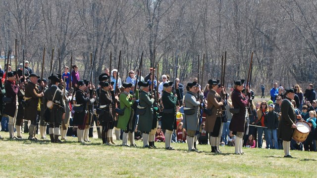 Colonial Militia stand in an open field and practice drilling with muskets in front of a large crown
