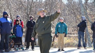 A National Park Ranger surrounded by visitors points off camera while speaking
