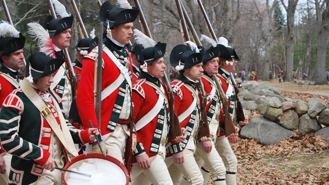 A group of 8 men dressed as Revolutionary War British soldiers with red coats, muskets and one drum.