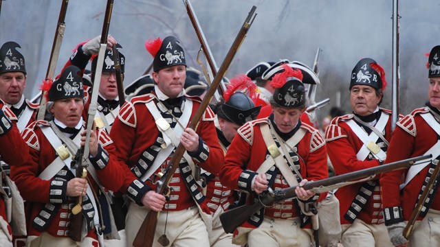 A group of red coated British soldiers advance forward with muskets at the ready