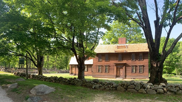 A wooden house surrounded by leafy trees. A stone wall is situated in front of the house.