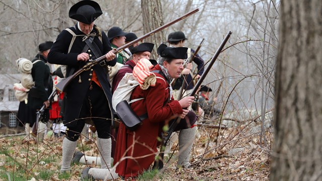 A group of militia soldiers prepare their muskets and look out over a stonewall.