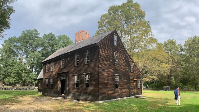 A wooden colonial house sits surrounded by trees. A person in colonial clothing stands in front