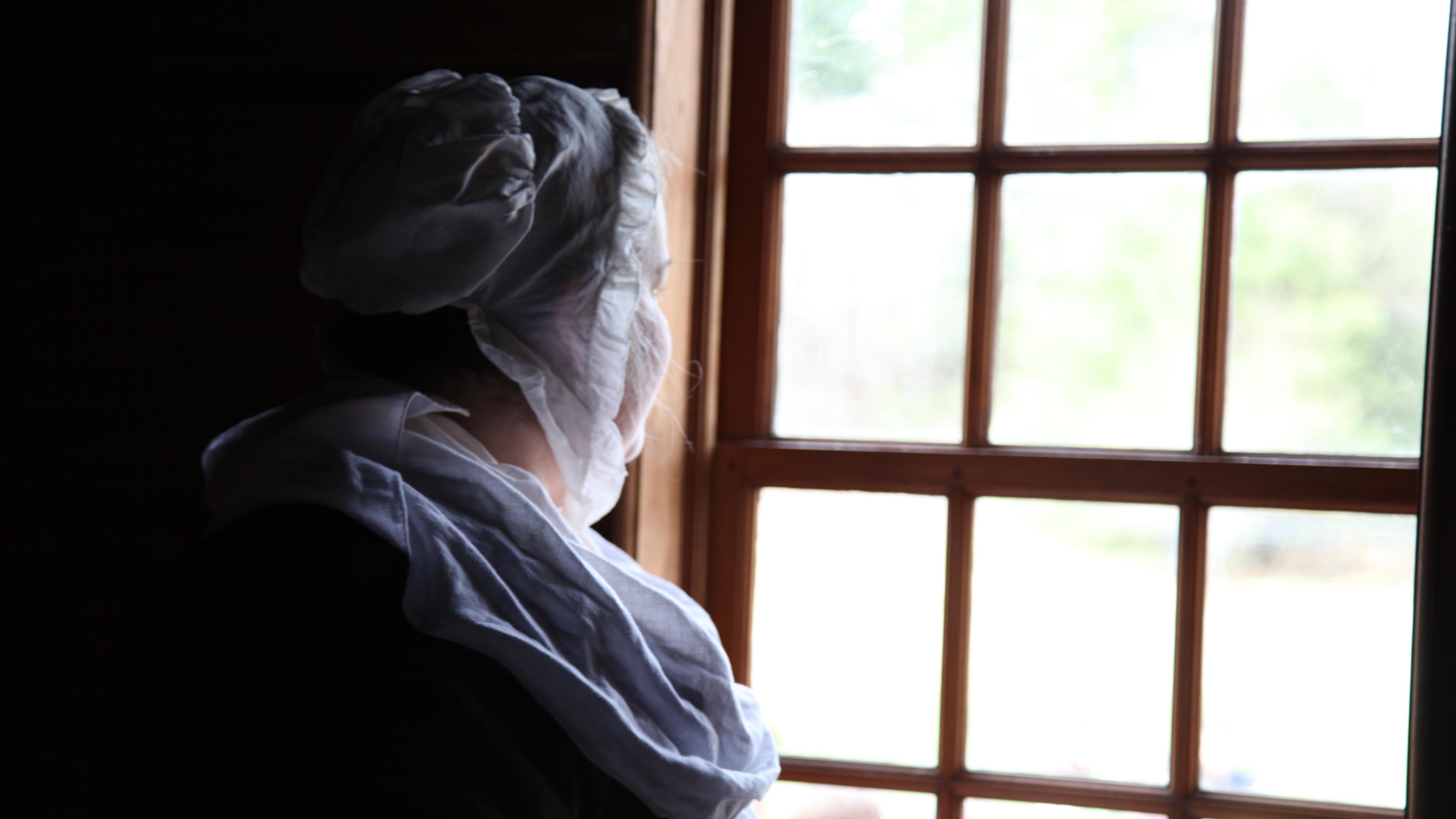 A woman in colonial dress with a white cap and neckerchief stares out a multi-paned window.