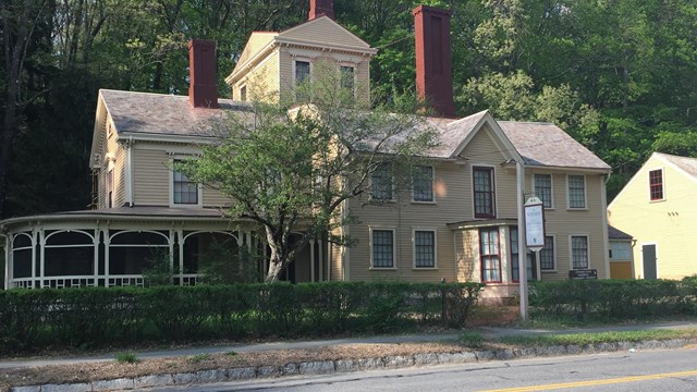 Large yellow house with a tower on the back viewed from across a street with a hedge in front