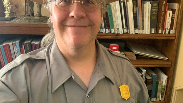 Man in NPS uniform and flat hat stands in front of a book shelf