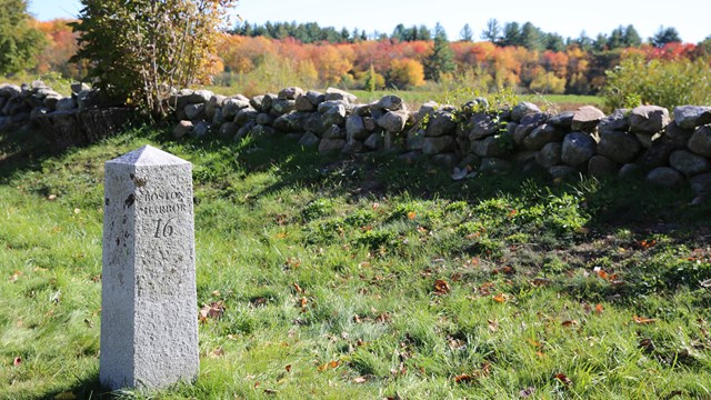 A stone mile marker reading "Boston 16 Miles" stands in a field with stonewall and fall trees behind