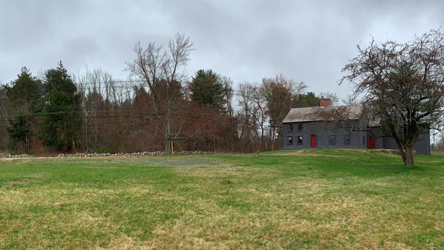 A large open field with a wooden house off to the right. A tall hill covered in trees in background
