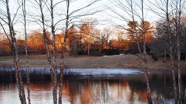 The Concord River is thinly frozen over with the sunrise turning the distant trees orange.